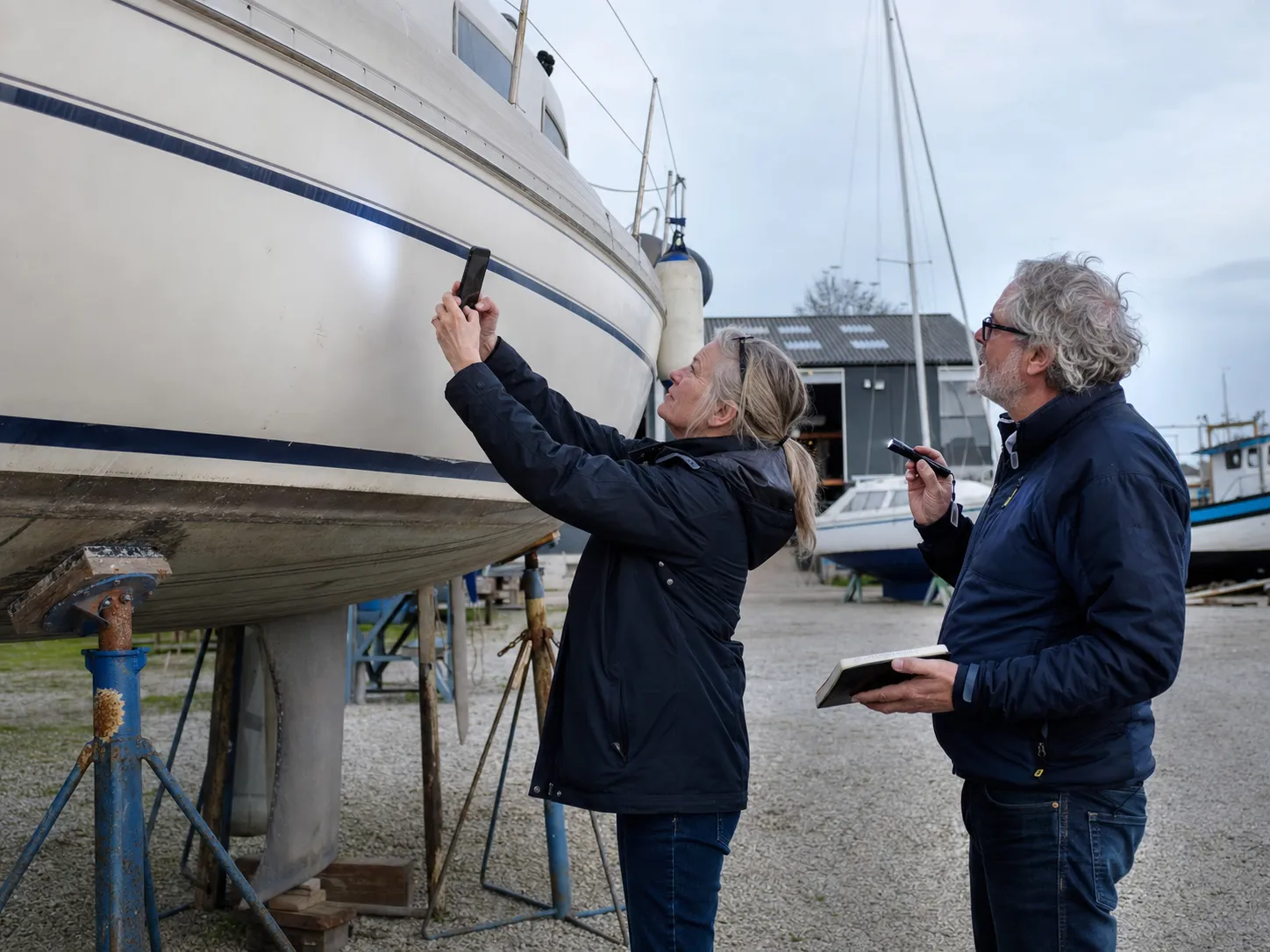 Eine Frau fotografiert bei einer Besichtigung den Rumpf eines Segelboots, während ihr Begleiter Notizen macht